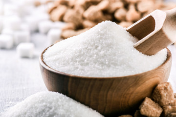 White sugar in wooden bowl with scoop on white table. White and brown sugar cubes in background.