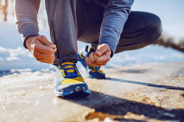 Close up of caucasian sporty man crouching outdoors in sportswear and tying shoelace. Winter fitness concept.