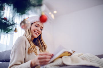 Beautiful caucasian young woman in sweater and with santa hat on head sitting on sofa next to...