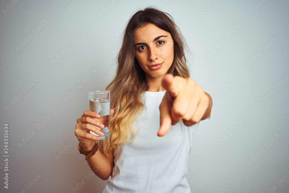 Sticker Young beautiful woman drinking a glass of water over white isolated background pointing with finger to the camera and to you, hand sign, positive and confident gesture from the front