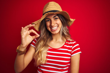 Young beautiful woman wearing summer hat over red isolated background doing ok sign with fingers,...