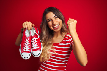 Young beautiful woman holding sneakers over red isolated background screaming proud and celebrating...