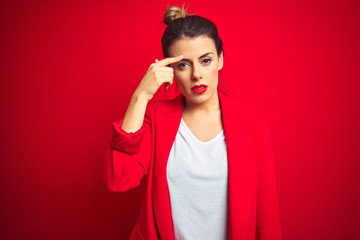 Young beautiful business woman standing over red isolated background pointing unhappy to pimple on...