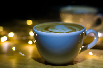 Fototapeta premium A white ceramic cup of cappuccino with latte art amid yellow flashlights in a dark coffee shop. Garland. Holiday.