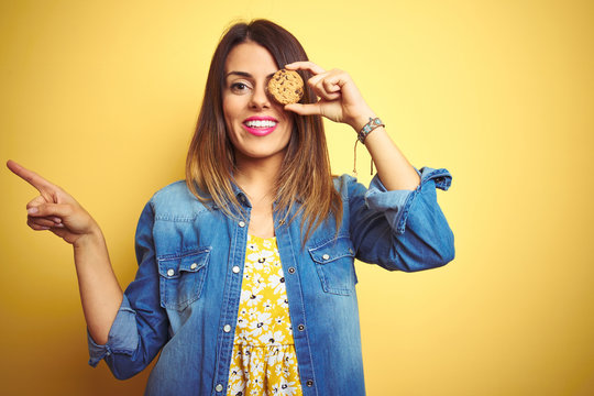 Young beautiful woman eating chocolate chips cookie over yellow background very happy pointing with hand and finger to the side
