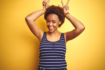 Beauitul african american woman wearing summer t-shirt over isolated yellow background Posing funny and crazy with fingers on head as bunny ears, smiling cheerful