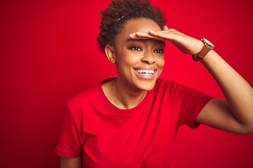 Young beautiful african american woman with afro hair over isolated red background very happy and smiling looking far away with hand over head. Searching concept.