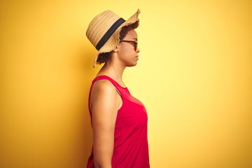 African american woman wearing summer hat and sunglasses over yellow isolated background looking to side, relax profile pose with natural face with confident smile.
