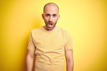 Young bald man with beard wearing casual striped t-shirt over yellow isolated background In shock face, looking skeptical and sarcastic, surprised with open mouth