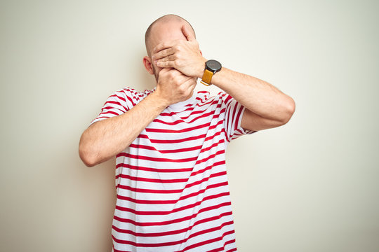 Young bald man with beard wearing casual striped red t-shirt over white isolated background Covering eyes and mouth with hands, surprised and shocked. Hiding emotion