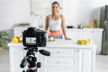 selective focus of digital camera with happy woman gesturing near vegetables and fruits on screen