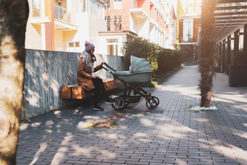 Young mother taking break by sitting on bench in urban living block. Woman walking with baby stroller down city streets.