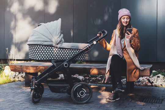 Woman walking with child in baby stroller and having rest in recreation area. Girl watching video online on screen of smartphone.