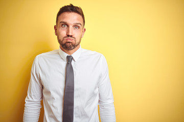 Young handsome business man wearing elegant white shirt over yellow isolated background puffing cheeks with funny face. Mouth inflated with air, crazy expression.