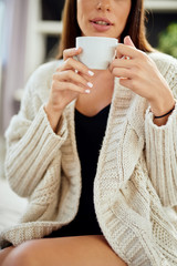 Caucasian woman dressed in beige sweater sitting on bed and holding fresh morning coffee.