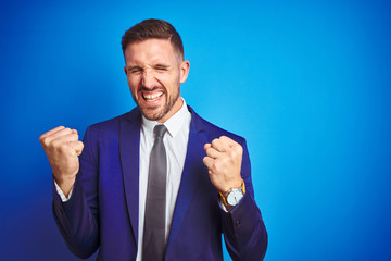 Young handsome business man over blue isolated background very happy and excited doing winner gesture with arms raised, smiling and screaming for success. Celebration concept.