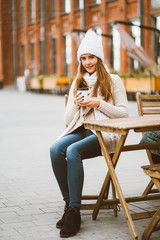 Beautiful young girl drinking coffee, tea from plastic reusable mug in autumn, winter. Woman with long hair, in warm clothes sitting in street cafe, warmed by hot drink, vertical