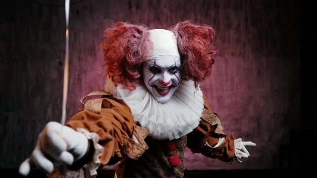 Creepy clown with colorful makeup in carnival costume looking at the camera suggests taking a red balloon. Frightening clown smiles ominously on dark isolated background. Slow motion shooting