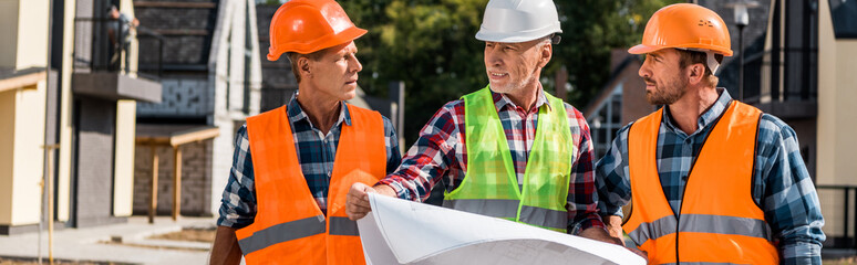 panoramic shot of mature builder holding blueprint near coworkers