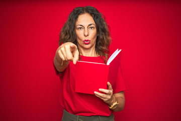 Middle age senior woman reading a book over red isolated background pointing with finger to the camera and to you, hand sign, positive and confident gesture from the front