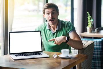 Young shocked businessman in green t-shirt sitting, pointing and showing laptop display. surprised face and looking at camera. business and freelancing concept. indoor shot near big window at daytime.