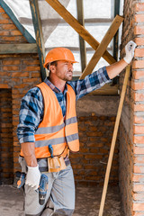 handsome constructor in helmet holding hammer drill while standing near brick wall