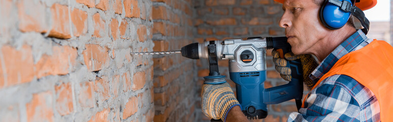 panoramic shot of mature builder using hammer drill near brick wall