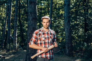 Naklejka na ściany i meble A handsome young man with a beard carries a tree. Stylish young man posing like lumberjack. Lumberjack worker walking in the forest with axe.