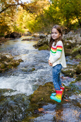 Naklejka na ściany i meble Outdoor recreation and awesome adventures with kids. A little child girl is walking along a green river in the forest in rubber boots on a warm autumn day. exploring nature, travel, family vacation.