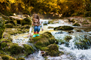 Outdoor recreation and awesome adventures with kids. A little child girl is walking along a green river in the forest in rubber boots on a warm autumn day.  exploring nature, travel, family vacation.