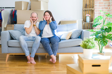 Fototapeta na wymiar Young couple sitting on the sofa arround cardboard boxes moving to a new house covering ears with fingers with annoyed expression for the noise of loud music. Deaf concept.