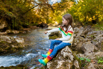 Outdoor recreation and  adventures with kids in fall. Little child girl sitting on stone near the river in the forest in rubber boots on warm autumn day. exploring nature, travel, family vacation.