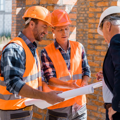 selective focus of constructors near mature businessman in helmet looking at blueprint