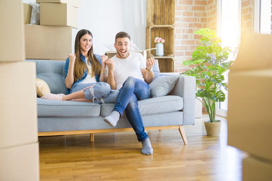Young beautiful couple relaxing sitting on the sofa around boxes from moving to new house celebrating surprised and amazed for success with arms raised and open eyes. Winner concept.