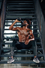 Fototapeta na wymiar Fit shirtless Black man sitting on metal steps and drinking fresh water from the bottle after working out