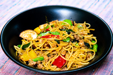 Tasty Thai spicy stir-fried spaghetti with pork, hot basil, green, yellow and red peppers in black bowl on wooden table.