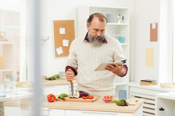 Waist up portrait of bearded senior man looking up recipe via digital tablet while cooking healthy vegetables in kitchen, copy space