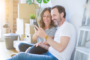 Middle age senior romantic couple in love sitting on the apartment floor with boxes around and using smartphone smiling happy for moving to a new home