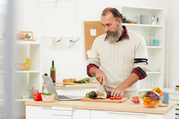 Waist up portrait of bearded senior man watching video recipe via laptop while cooking healthy salad in kitchen, copy space