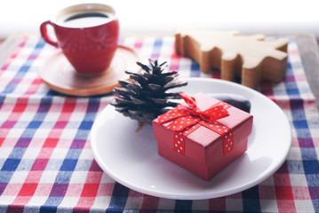 Close up red gift box decorate with pine cones on white dish lay on napery with red coffee cup and christmas tree on background