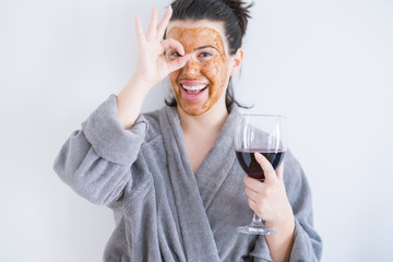 Beautiful woman wearing cosmetic facial mask as skincare treatment drinking glass of wine with happy face smiling doing ok sign with hand on eye looking through fingers