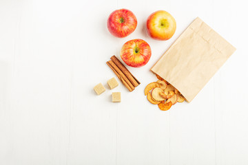 Organic homemade dry fruit chips in a paper eco pack and fresh apples, cinnamon sticks, cane sugar on a white background. Healthy vegan snack of apples. Ingredient for baking, cooking.
