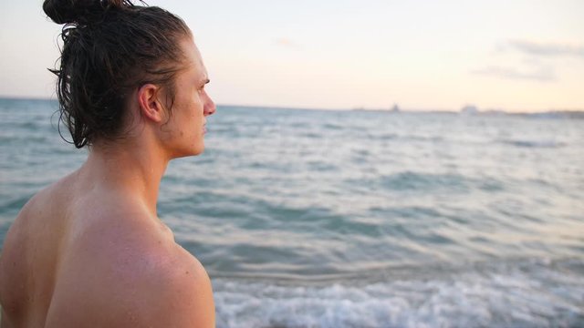 Young Man on Beach looking at Spanish Sea Waves. Long Haired Surfer Guy at Blue Sky Ocean on a Summer Holiday Palm Tree Beach in Spain.