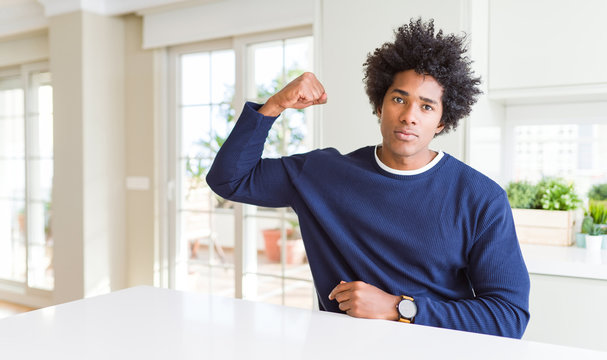 Young african american man wearing casual sweater sitting at home Strong person showing arm muscle, confident and proud of power