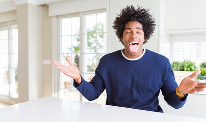 Young african american man wearing casual sweater sitting at home sticking tongue out happy with funny expression. Emotion concept.