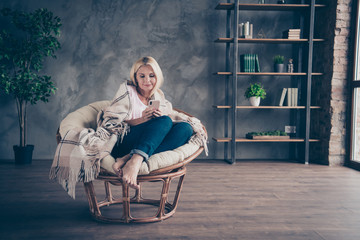 Full size photo of focused middle aged blonde hair woman use her cellphone read social media network enjoy autumn holidays sit on furniture white armchair covered with checkered blanket in living room