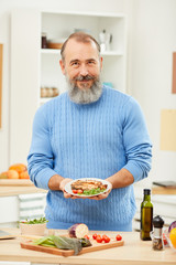 Waist up portrait of bearded senior man presenting meat steak and smiling at camera while cooking in kitchen at home