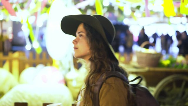 Elegant woman in hat walking on traditional easter fair in downtown of Prague at night. Side view young girl drinking mulled wine, enjoying decorated fairgrounds against illuminated stalls 