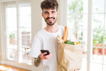 Young handsome man holding a paper bag full of fresh groceries at home, showing and using smartphone while buying products