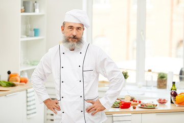 Waist up portrait of bearded senior chef looking at camera while posing confidently standing in restaurant kitchen, copy space